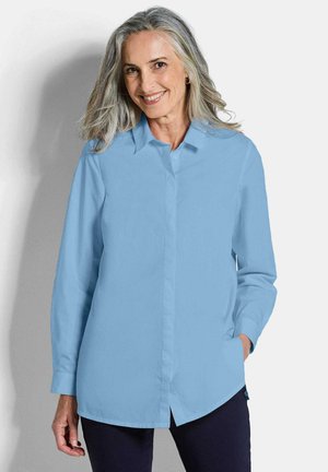 Femme souriante aux cheveux gris portant une chemise bleu clair boutonnée et un pantalon foncé, debout avec une main dans la poche devant un fond uni.