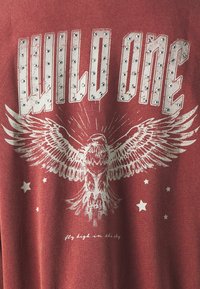 T-shirt rosso mélange con la scritta "WILD ONE" in bianco e dettagli in applicazione. Un'illustrazione di un'aquila con le ali spiegate e stelle, con il testo sottostante che recita "vola in alto nel cielo."
