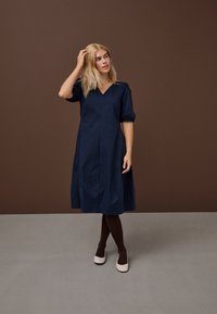 Ženska s svetlimi lasmi, oblečena v mornarsko modro srednje dolgo obleko, temne najlonke in bež balerinke, stoji pred rjavim in sivim ozadjem.