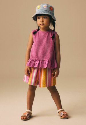 Young girl with braided hair wearing a gray hat with flower patches, sleeveless purple top, striped shorts, and floral sandals standing against beige background.