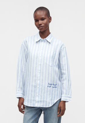 Persona che indossa una camicia a righe azzurro chiaro e bianco con bottoni, con il testo "YOUR BEST BAD IDEA" ricamato vicino all'orlo, e jeans azzurri.