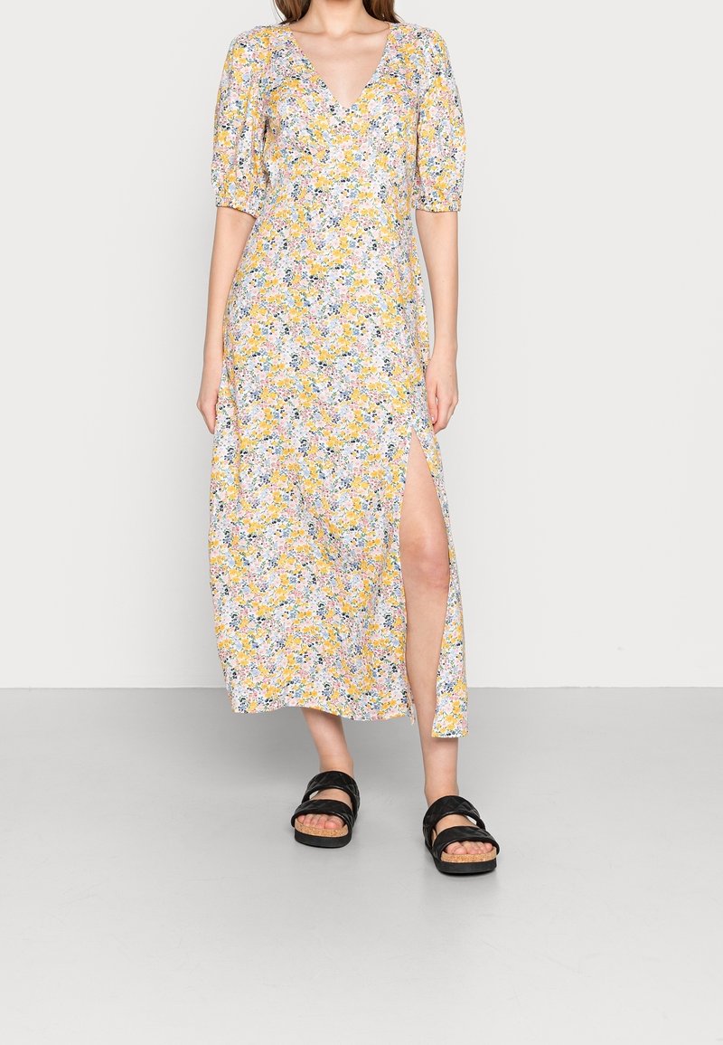 Robe maxi florale avec un imprimé jaune, rose et vert, décolleté en V, manches bouffantes et fente latérale. Portée avec des sandales à lanières noires.