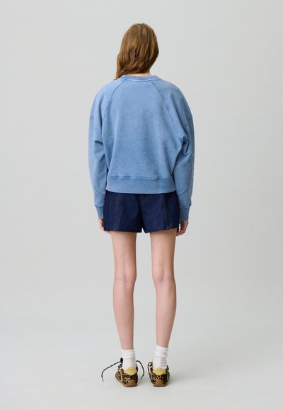 Felpa azzurra con maniche raglan, abbinata a pantaloni corti di jeans scuri. Le calzature presentano una stampa leopardata. Sfondo semplice.