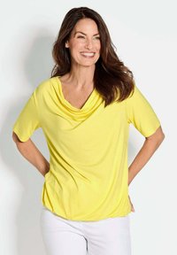 Geel gedrapeerd T-shirt met korte mouwen, zachte stof, losse pasvorm, gedragen met witte broeken. Gladde textuur, minimalistisch ontwerp, casual uitstraling.