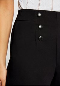 Pantalon noir taille haute avec trois boutons noirs mats, tissu lisse et silhouette ajustée avec poches latérales.