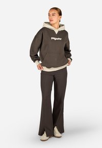 Hnedá mikina s kapucňou a béžovou podšívkou, doplnená tmavohnedými rozšírenými nohavicami so bočnými rozparkami a béžovými teniskami.