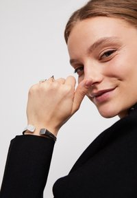 Pulsera de plata con acentos cuadrados sobre una banda negra, llevada en una mano. La ropa oscura y un fondo neutro resaltan los accesorios.