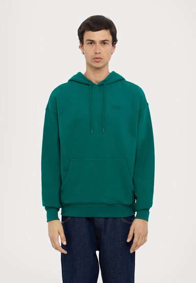 Green cotton hoodie featuring a front pocket, drawstrings, and a subtle logo. Ribbed cuffs and hem; relaxed fit. Worn with dark jeans.