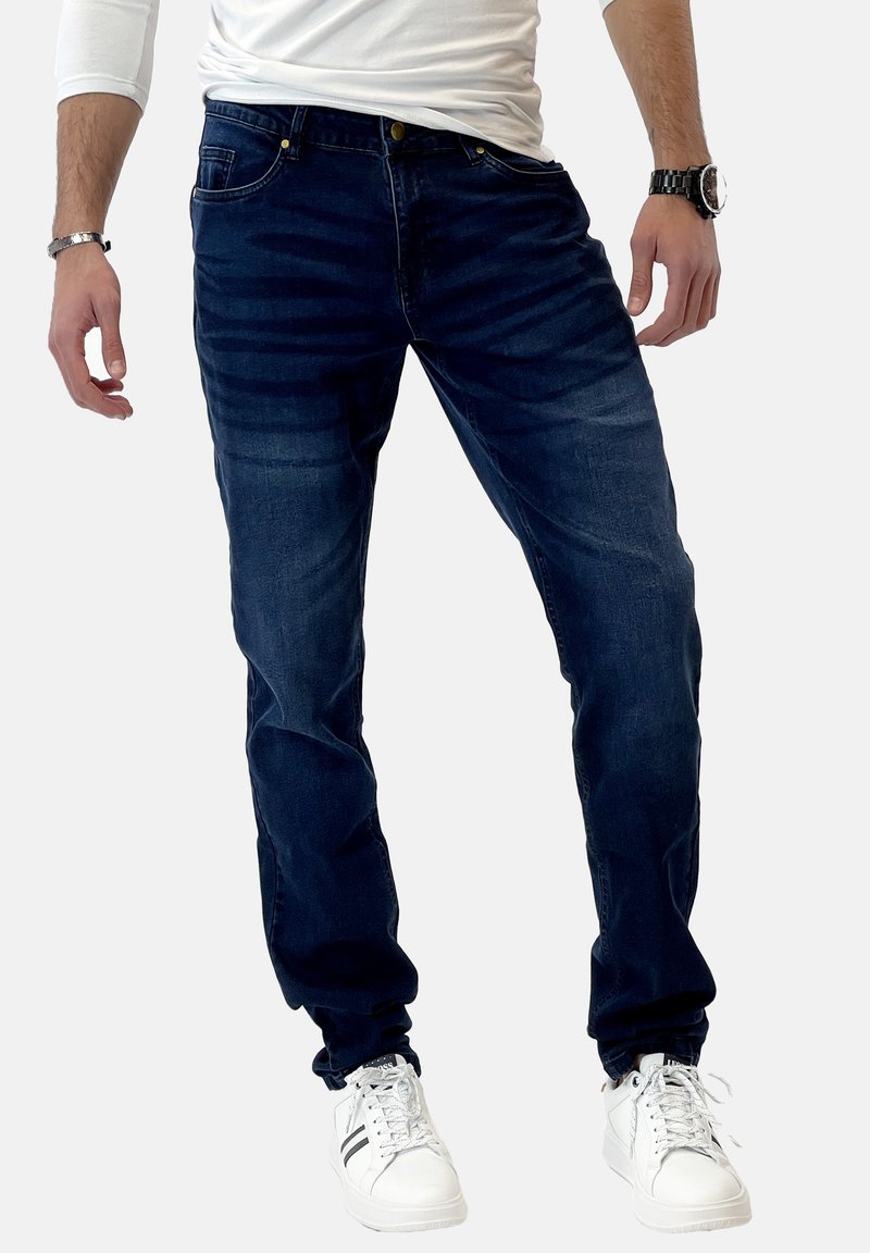 Jeans in denim blu scuro con un design aderente, dettagli di sbiadimento delicati, cinque tasche e bottone dorato, abbinati a sneakers bianche.