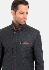 Steppjacke aus schwarzem Leder mit hohem Kragen, strukturiertem Stoff, braunen Akzenten und einer mit Reißverschluss versehenen Brusttasche.