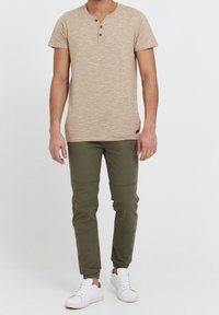 Beige kortärmad henley-tröja med tre bruna knappar, i kombination med olivgröna smalrandiga joggerbyxor och vita sneakers.