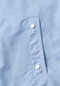 Tela azul claro con un bolsillo vertical asegurado por dos botones a presión plateados, uno cerca de la parte superior y otro cerca de la parte inferior de la solapa del bolsillo.