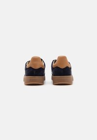 Navy suede sneakers with brown leather accents, textured rubber soles, and contrasting stitching. Features a rounded heel and lace holes.