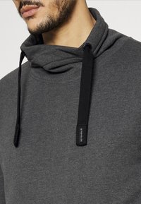 Sweat à capuche gris doté d'une texture douce, d'un col bénitier et de cordons noirs réglables. Comprend une petite étiquette logo sur le côté.