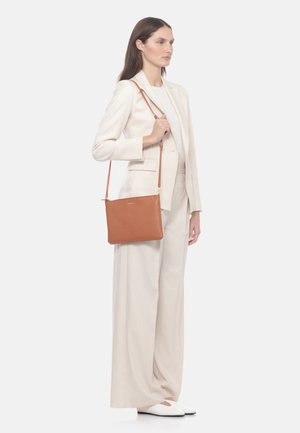 Femme en costume beige clair porte un sac en cuir marron moyen à bandoulière fine, debout devant un fond clair uni.