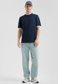 Granatowa koszulka z asymetrycznym szwem, zestawiona z jasnoniebieskimi dżinsami. Model nosi jasnoniebieską czapkę i szare trampki z białymi akcentami.