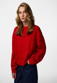 Red cable-knit sweater with a relaxed fit, round neckline, and ribbed cuffs. Worn with dark denim pants. Minimalist background.