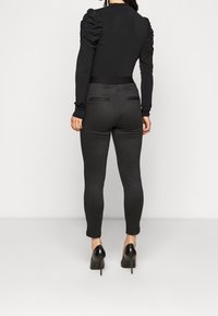 Svart långärmad topp med puffade axlar, kombinerad med figurnära mörkgrå rutiga byxor och svarta stilettoklackar, sedd bakifrån.