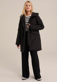 Parka impermeabil negru cu glugă, închidere prin fermoar și butoane, buzunare frontale și croială relaxată. Purtați peste o cămașă în dungi și pantaloni negri.