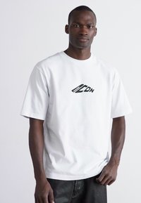 T-shirt en coton blanc avec un design de logo noir sur le devant. Manches courtes et coupe décontractée. Porte avec un pantalon noir.