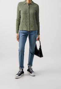 Camisa de botones texturizada en verde oliva, jeans azules, zapatillas negras y un pequeño bolso negro. La camisa tiene un cuello y siete botones.