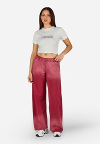 Izgriezta balta T-kreklā ar uzrakstu "AUKSTAS ROkas SILTA SIRDS", kopā ar rozā platām biksēm, kurām ir elastīga josta un gluda auduma tekstūra.