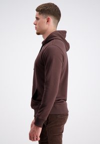 Brauner Hoodie aus weichem Stoff, mit einer Kängurutasche, Kapuze mit Kordelzug und gerippten Bündchen. Getragen mit dunkelbraunen Hosen.