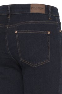 Tmavě modré džíny z denimu, pohled zezadu ukazující pravou kapsu s měděnými nýty, kontrastním šitím a hnědou koženou nášivkou značky „PULZ JEANS".