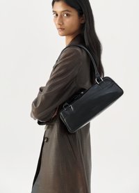 Bolso de cuero negro con forma rectangular y asas cortas, que presenta una textura suave y un branding sutil, llevado sobre un abrigo marrón texturizado.