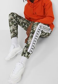 Personne portant un sweat à capuche orange, un pantalon de survêtement camouflage avec des bandes blanches à logo, des chaussettes blanches et des baskets blanches assise par terre.