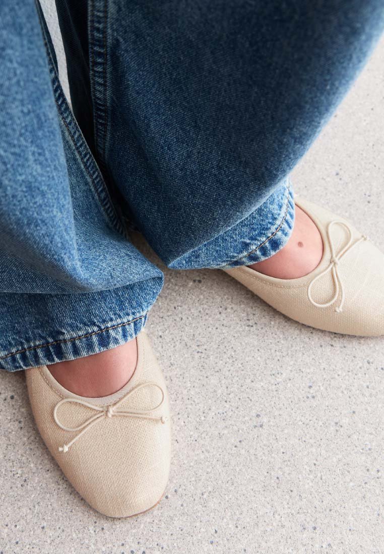 Bailarinas de lona beige con detalle de lazo, combinadas con jeans de pierna ancha azul, vistas desde arriba sobre un suelo claro con manchas.