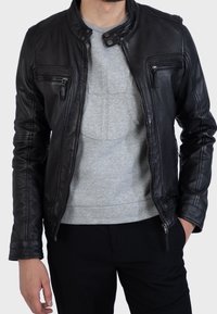 Musta nahkatakki, jossa on istuva malli, pystykaulus, vetoketjulliset taskut ja hienovaraiset ompelu-yksityiskohdat. Päällä harmaa paita.