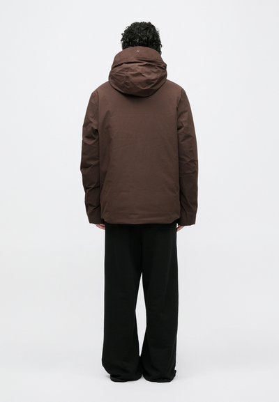 Brūna kapuci jaka no teksturēta materiāla, ar brīvu piegriezumu, redzamām šuvēm un kapuci ar auklu. Melnas plata izmēra bikses.