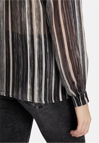 Gestreepte doorschijnende blouse in zwarte, bruine en beige tinten, gedragen met donkergrijze jeans, waarbij de manchet met zwarte knoop zichtbaar is en de hand naast het lichaam rust.