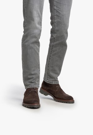 Bottines en suede marron avec une finition texturée, un design à lacets et une semelle en caoutchouc robuste. Portées avec un jean ajusté gris clair.