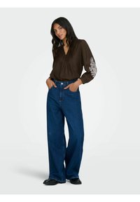 Brun langærmet bluse med v-udskæring og blomsterbroderi på ærmerne, parret med højtsiddende brede blå jeans. Sorte sko.