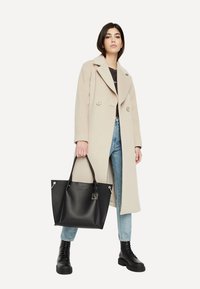 Schwarze Einkaufstasche aus strukturiertem Leder, mit silbernen Beschlägen und einem Logotag. Getragen zu einem grauen Oberteil, blauen Jeans und schwarzen Kampfstiefeln.