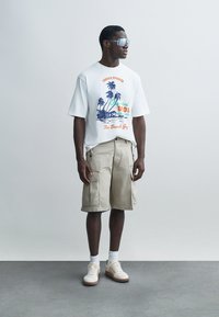 T-shirt en coton blanc avec imprimé tropical coloré, assorti à un short cargo beige et des baskets blanches avec des accents texturés et des semelles claires.