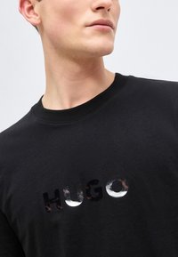 Νεαρός άνδρας που φοράει μαύρο μπλουζάκι με στρογγυλή λαιμόκοψη και λαμπερά μαύρα γράμματα "HUGO" στο στήθος, με απλό φόντο.