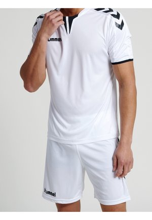 Athlet trägt weißes Hummel-Sporttrikot und Shorts mit schwarzen Akzenten, steht vor einem schlichten hellen Hintergrund.