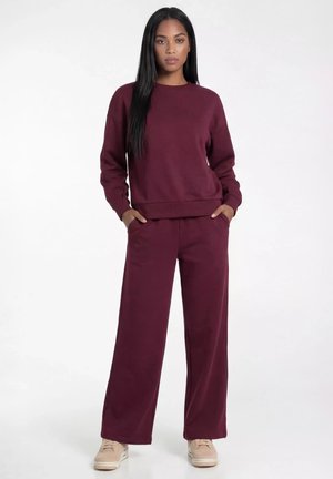 Kobieta z długimi czarnymi włosami, ubrana w dopasowaną bordową bluzę i szerokie spodnie, stoi z rękami w kieszeniach, ma na nogach beżowe trampki.