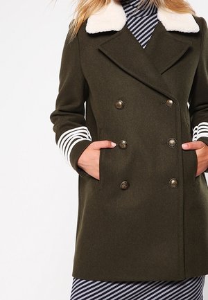 Femme portant un manteau vert foncé à double boutonnage avec un col en fausse fourrure blanche et des poignets rayés, les mains dans les poches au-dessus d'une robe rayée.