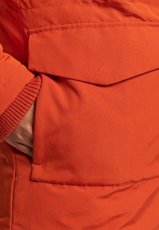 Hand gedeeltelijk binnenin een grote, flapbedekte zak van een oranje gewatteerde jas met een zichtbare geribbelde manchet.