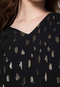 Schwarzes Stoffoberteil mit V-Ausschnitt, das verstreute metallische goldene Linien in verschiedenen Längen und Breiten aufweist. Glatte Textur.