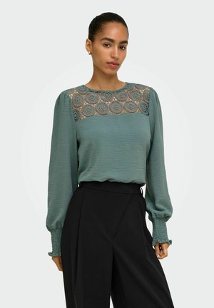 Femme portant un chemisier vert à manches longues avec un détail de dentelle florale sur le plastron et un pantalon noir taille haute, debout avec une expression neutre.