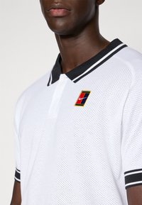 Hombre vistiendo una camiseta polo de malla blanca con cuello y puños negros, que presenta un parche rojo, azul y verde con el logo de Nike en blanco.