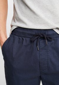 Personne portant un pantalon à cordon de serrage bleu marine avec taille élastique et un t-shirt décontracté gris, main glissée dans la poche gauche.