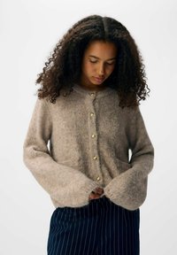 Brauner, flauschiger Strickcardigan mit Rundhalsausschnitt, langen Ärmeln und goldenen Knöpfen an der Vorderseite. Kombiniert mit navyblauen gestreiften Hosen.