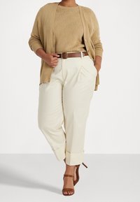 Nainen pukeutuneena beigeen neuletakkiin ja puseroon, kermaisiin korkeavyötäröisiin housuihin, joissa on käänteet, ruskeaan vyöhön ja ruskeisiin korkokenkiin.