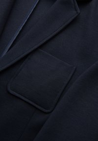 Dunkel marineblauer Mantelstoff mit einer rechteckigen Klappentasche und überlappenden Kragenrevers-Details in Nahaufnahme.